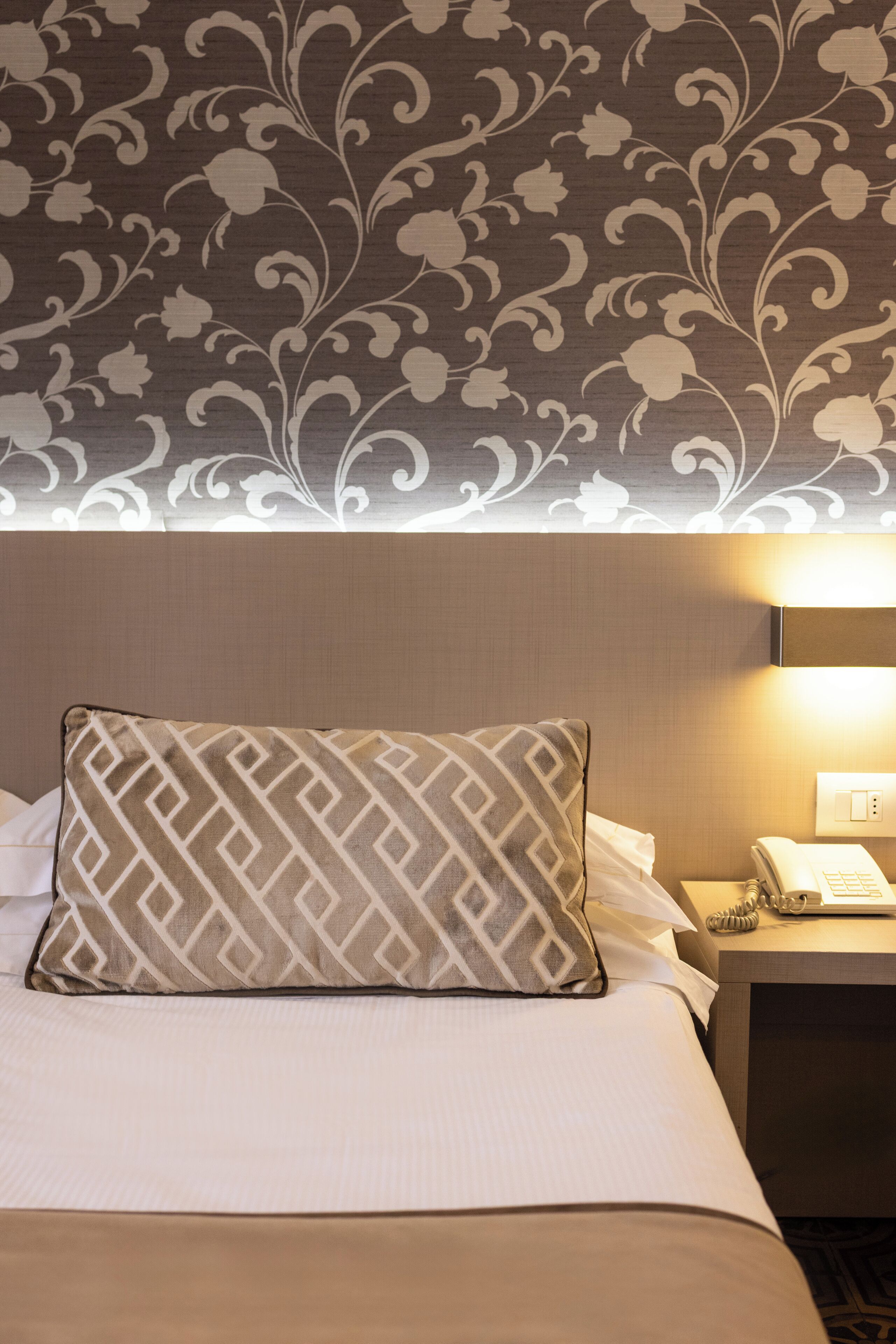 Habitación de hotel con papel tapiz estampado, cama con almohada estampada, mesita de noche con lámpara y teléfono.
