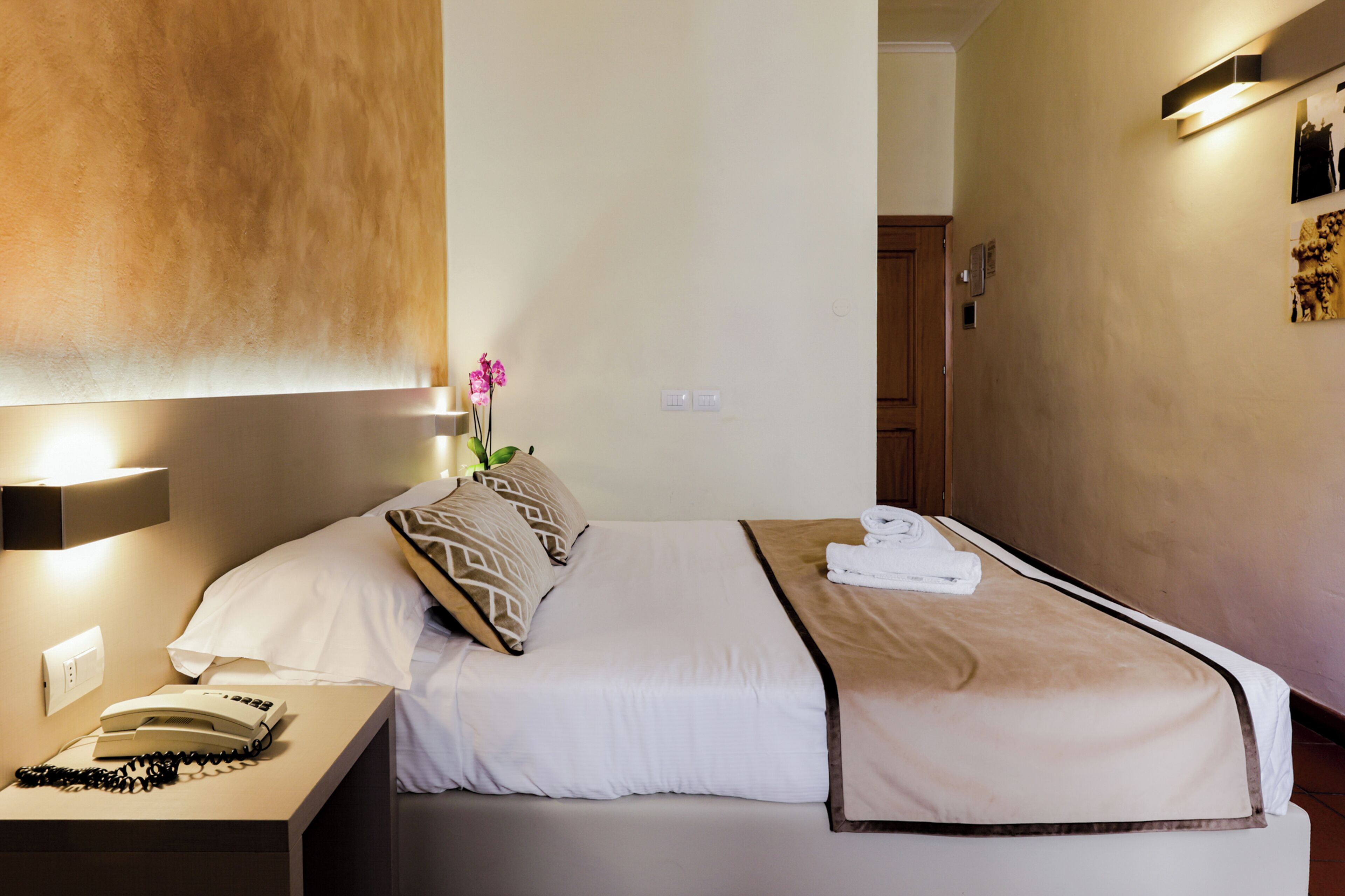 A hotel room with a neatly made bed, a bedside telephone, and a wall-mounted light fixture.