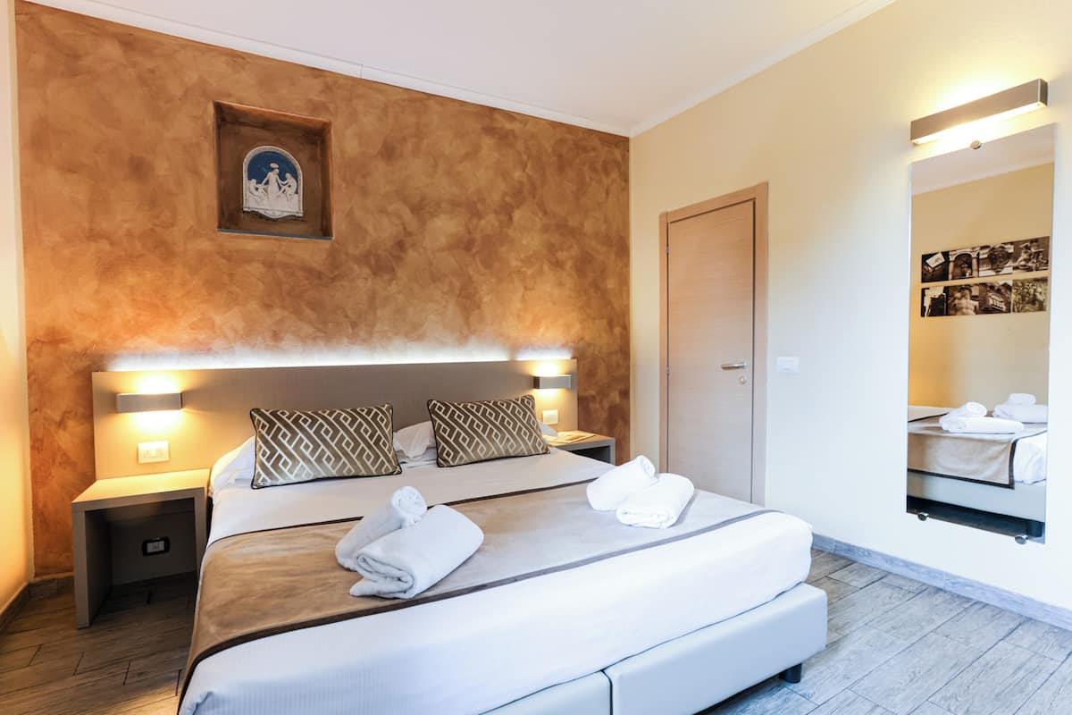 Une chambre d’hôtel avec un grand lit, deux tables de chevet, une petite table, une lampe fixée au mur, un tableau encadré et un miroir orné d’un photomontage.
