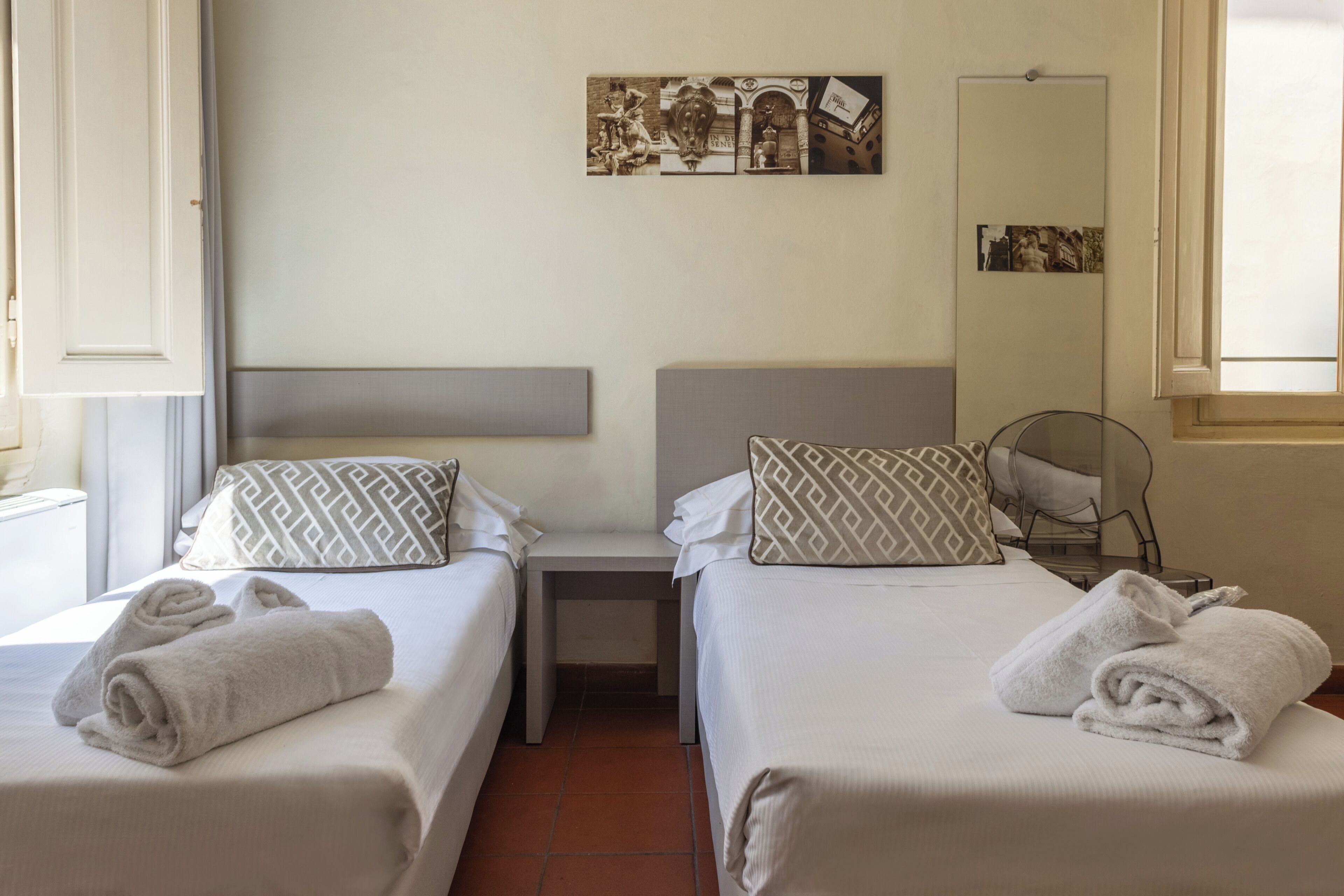 Une chambre d’hôtel avec deux lits, chacun muni d’un oreiller et d’une serviette roulée. Il y a une petite table entre les lits et un miroir au mur.