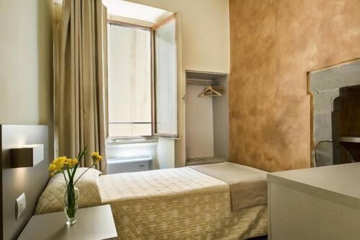 Une chambre d’hôtel comprenant un lit, une table de chevet avec un vase de fleurs, une fenêtre avec des rideaux, une armoire et un miroir.