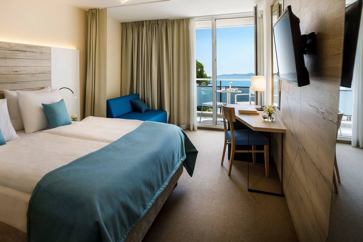 A hotel room with a large bed, a desk with a chair, a TV mounted on the wall, and a balcony with a view of the sea.
