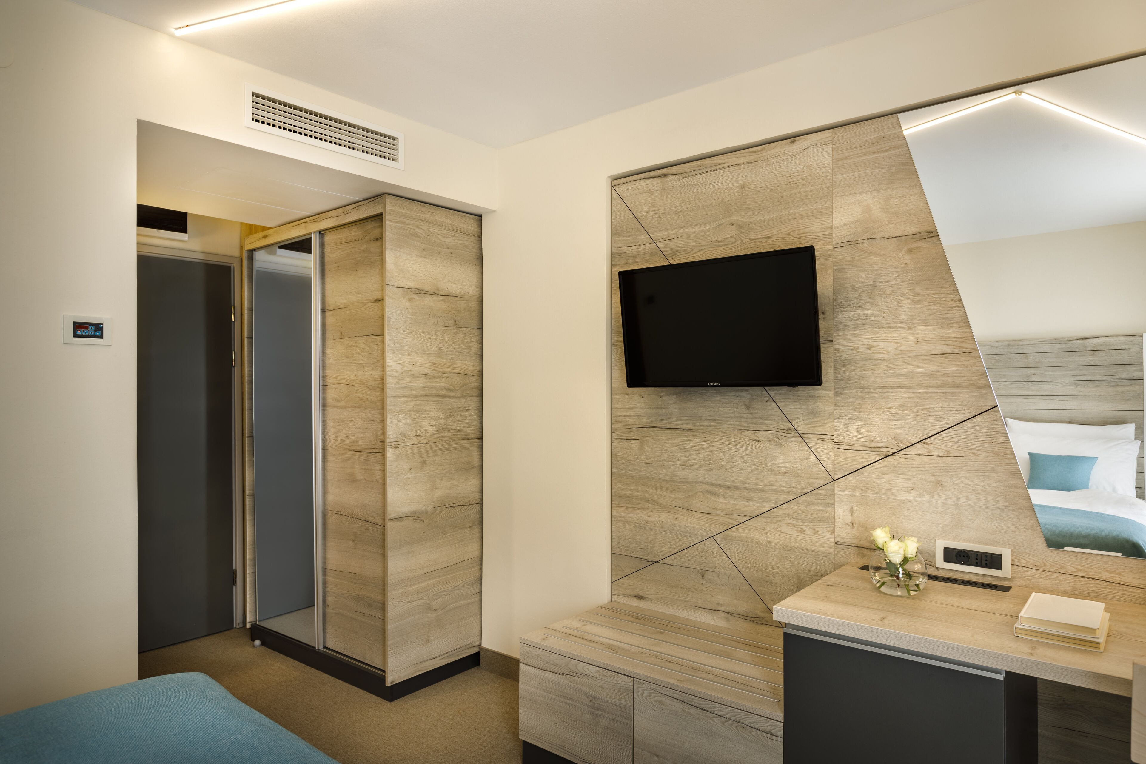 Ein modernes Hotelzimmer mit einer Wand im Holzdesign, einem flachbildfernseher über einem Schreibtisch, einem Nachttisch mit Blumen und einem großen Spiegel, der den Raum reflektiert.