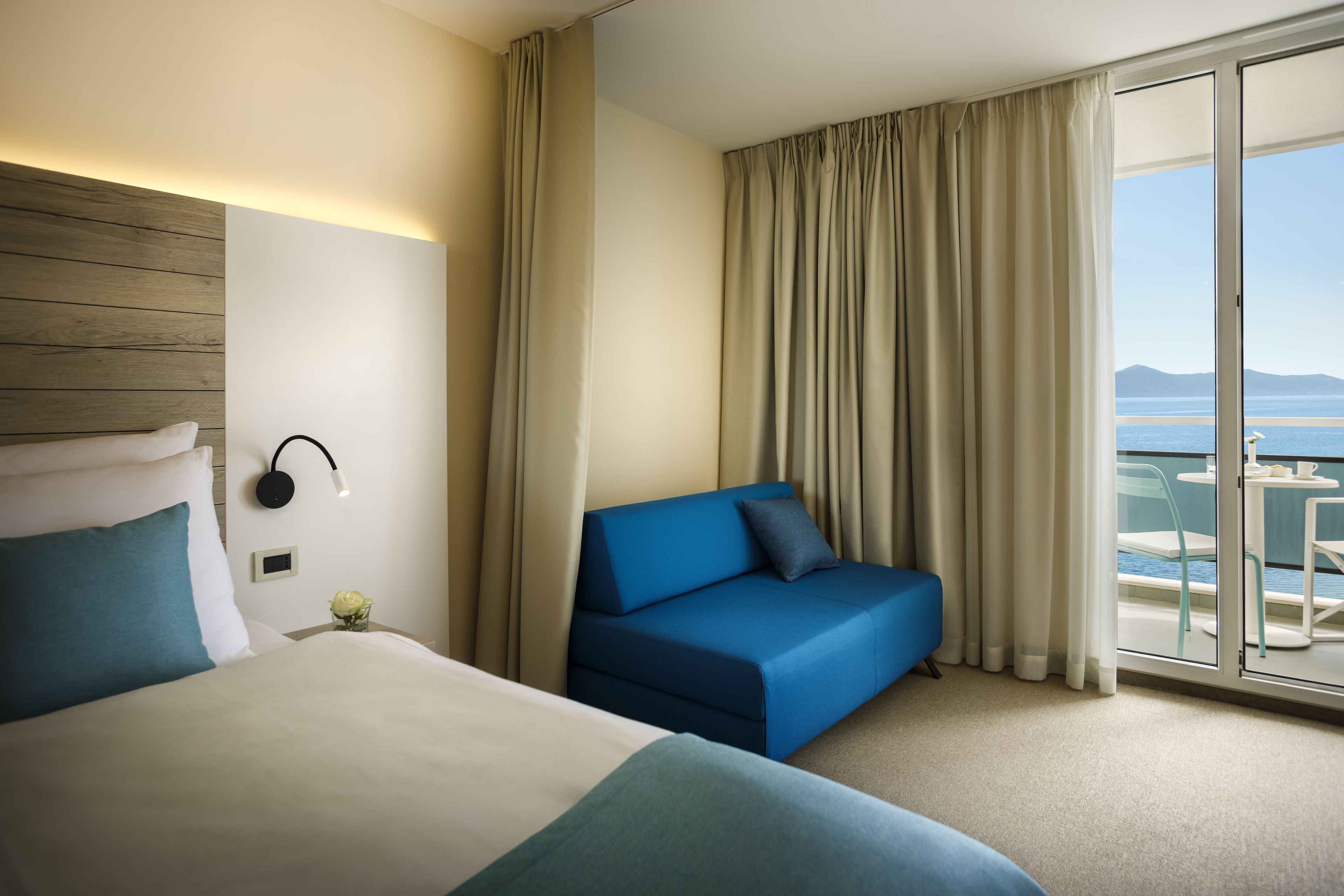 Habitación de hotel con cama, sofá azul, balcón y vista al mar, y cabecera de madera.