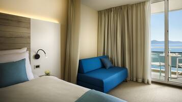 Habitación de hotel con cama, sofá azul, balcón y vista al mar, y cabecera de madera.