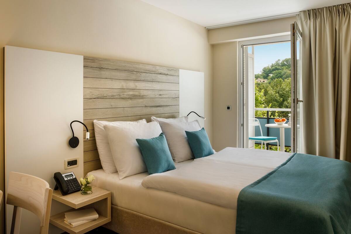 A hotel room with a bed, bedside table, phone, and a view of the outdoors.