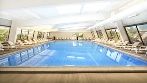 Una amplia piscina interior con agua cristalina, rodeada de tumbonas y grandes ventanales que ofrecen vistas a la vegetación.