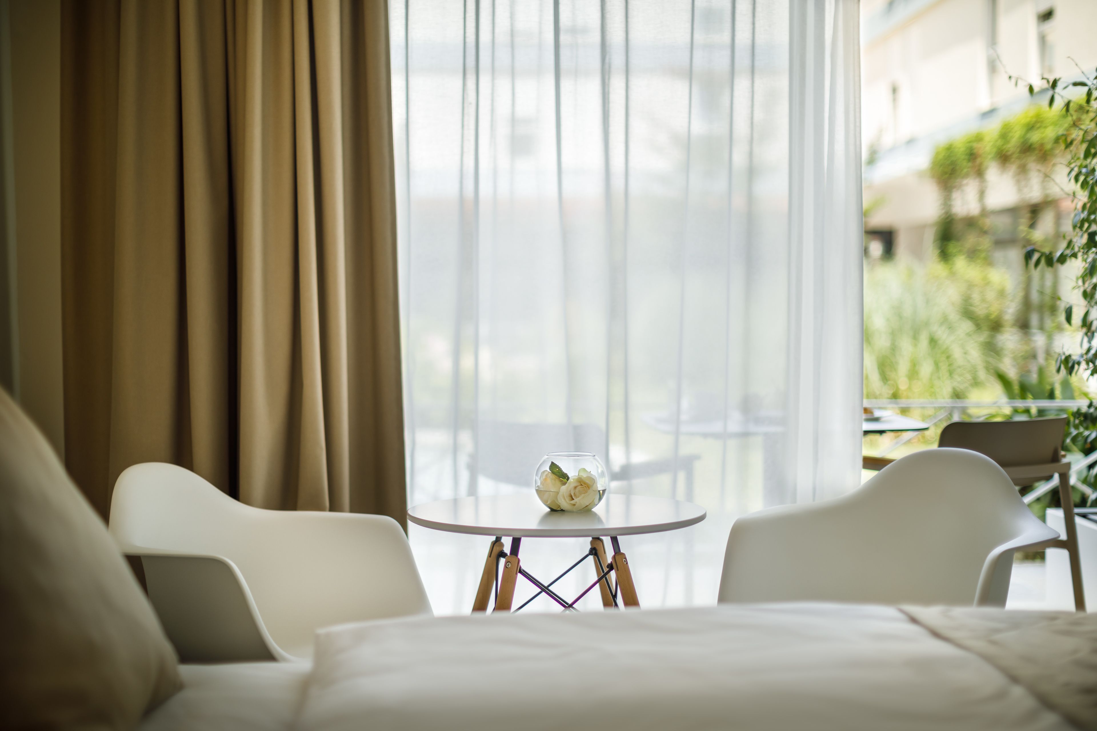 A modern hotel room with a bed, two white chairs, a small round table with a glass bowl of flowers, and a large window with curtains.