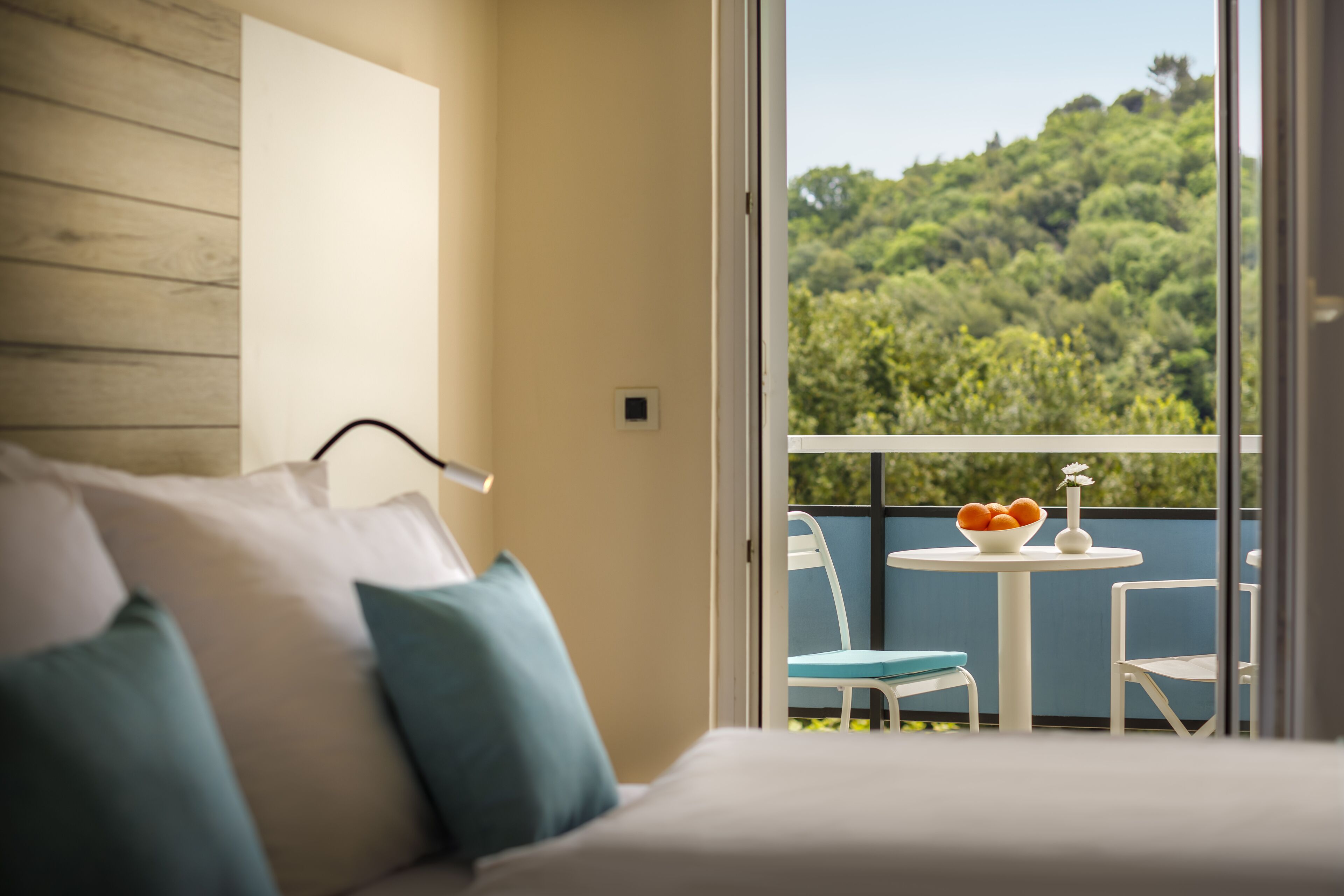 A hotel room with a bed, a small table with a bowl of fruit, and a view of a lush green landscape through large windows.
