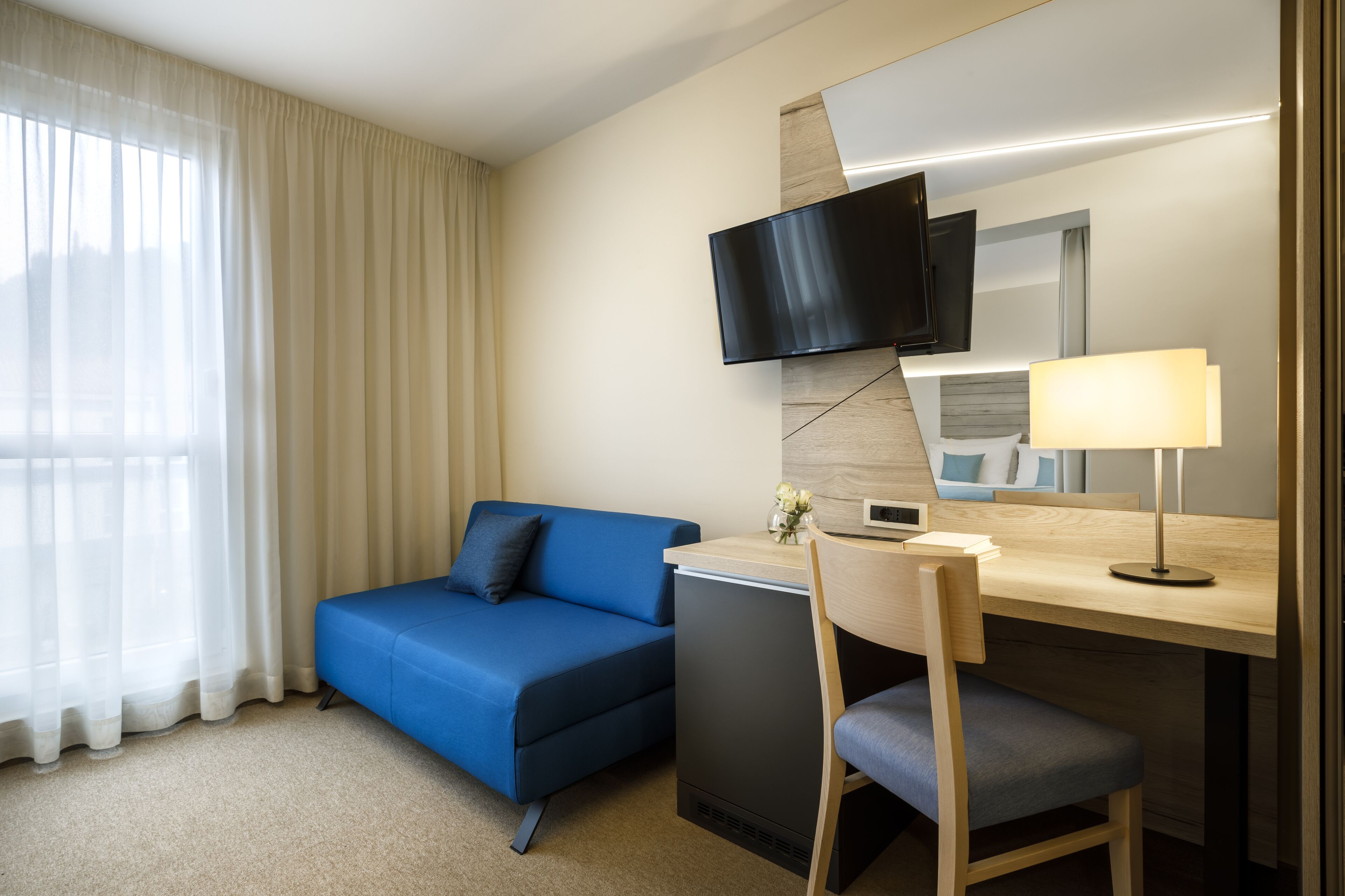 A hotel room with a blue sofa, a desk with a lamp, a flat-screen TV, and a small refrigerator.