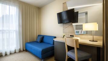 A hotel room with a blue sofa, a desk with a lamp, a flat-screen TV, and a small refrigerator.
