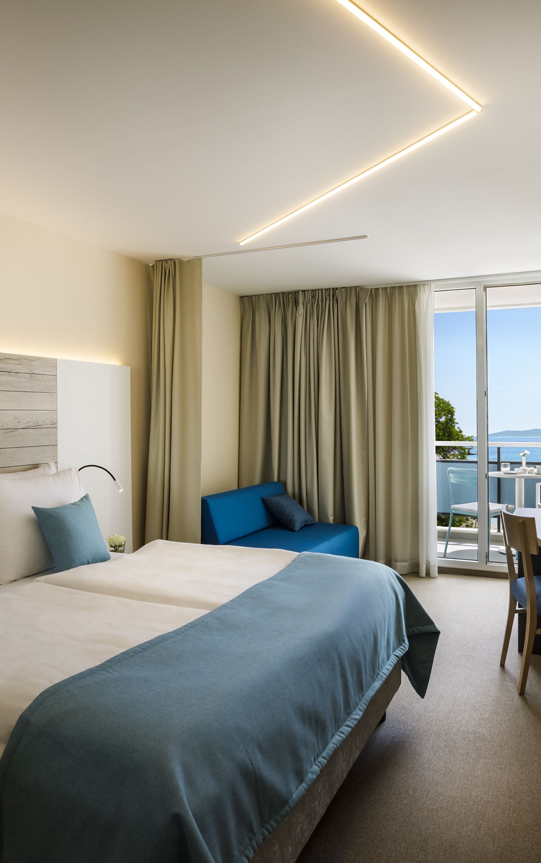 A modern hotel room with a large bed, a blue sofa, a balcony with a view, and a ceiling light fixture.