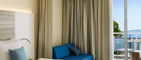 A modern hotel room with a large bed, a blue sofa, a balcony with a view, and a ceiling light fixture.