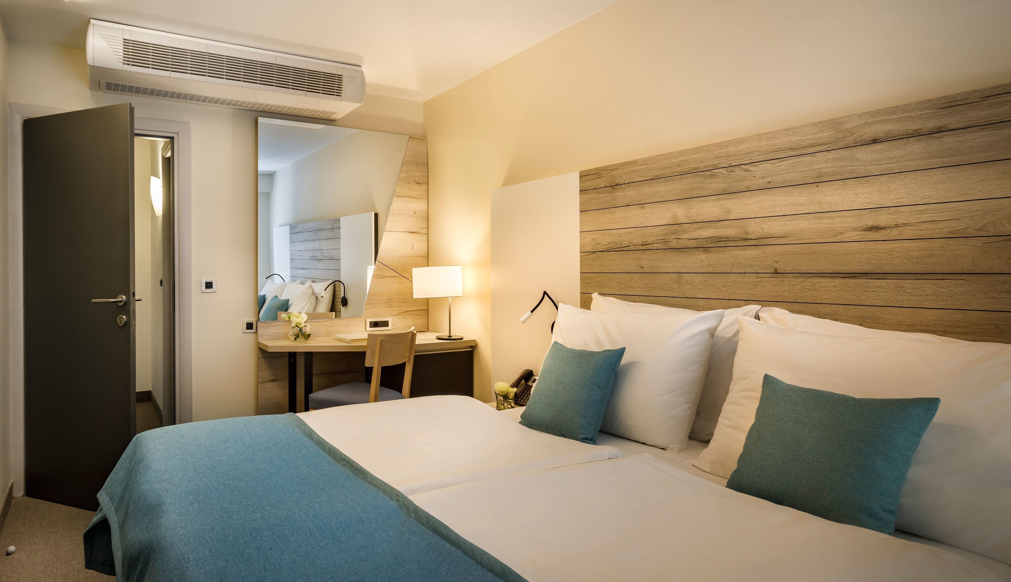 Quarto de hotel com cama, mesa de cabeceira, abajur e cabeceira de madeira.