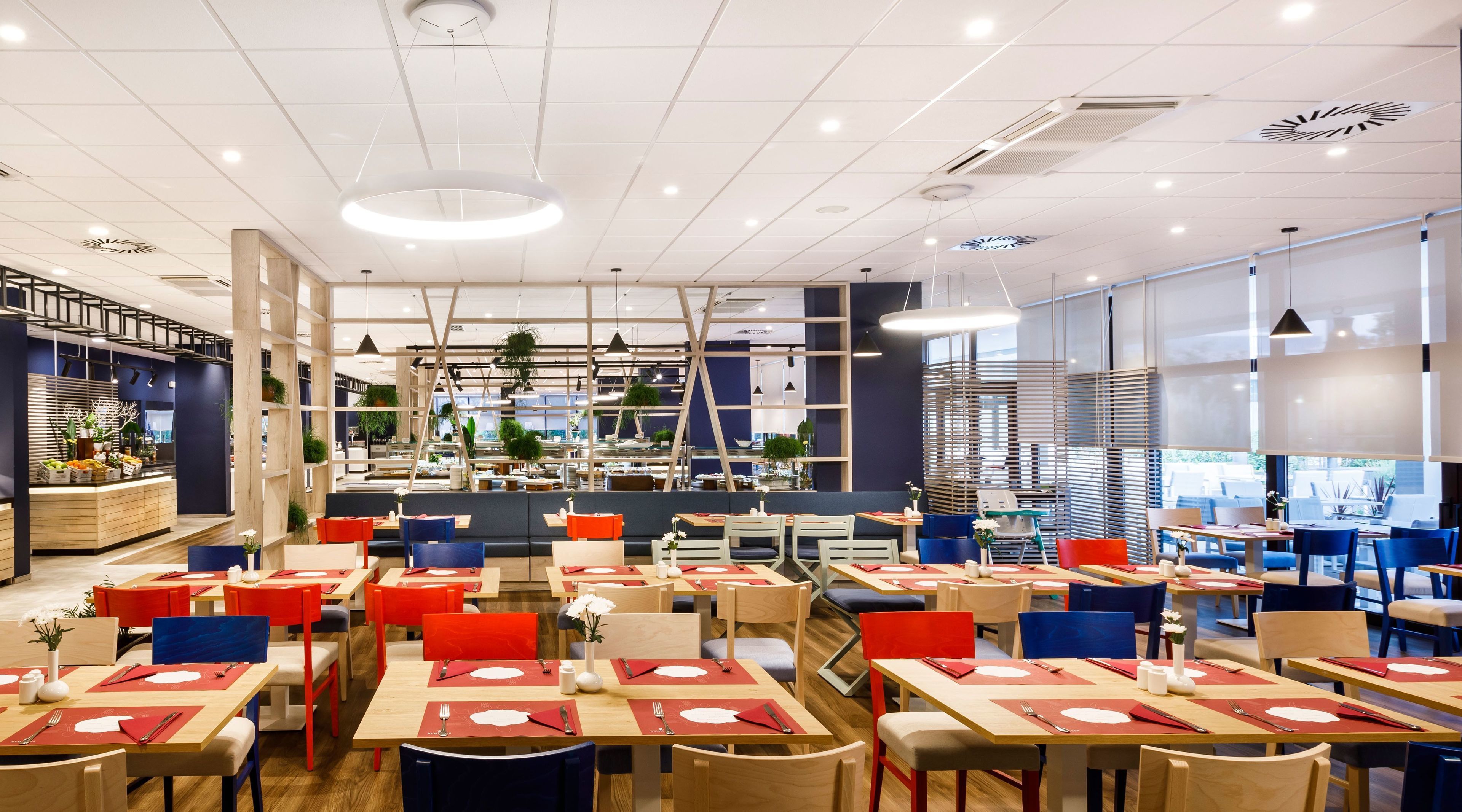 A modern restaurant interior with multiple dining areas, wooden tables, red and blue chairs, and a central seating area with a staircase.
