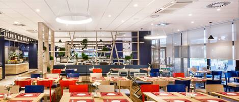A modern restaurant interior with multiple dining areas, wooden tables, red and blue chairs, and a central seating area with a staircase.