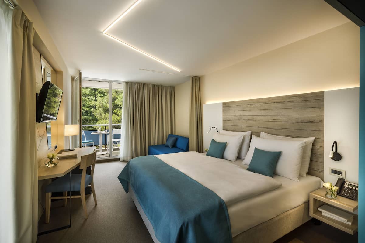 A hotel room with a large bed, a desk with a chair, a small table, and a view of greenery outside.