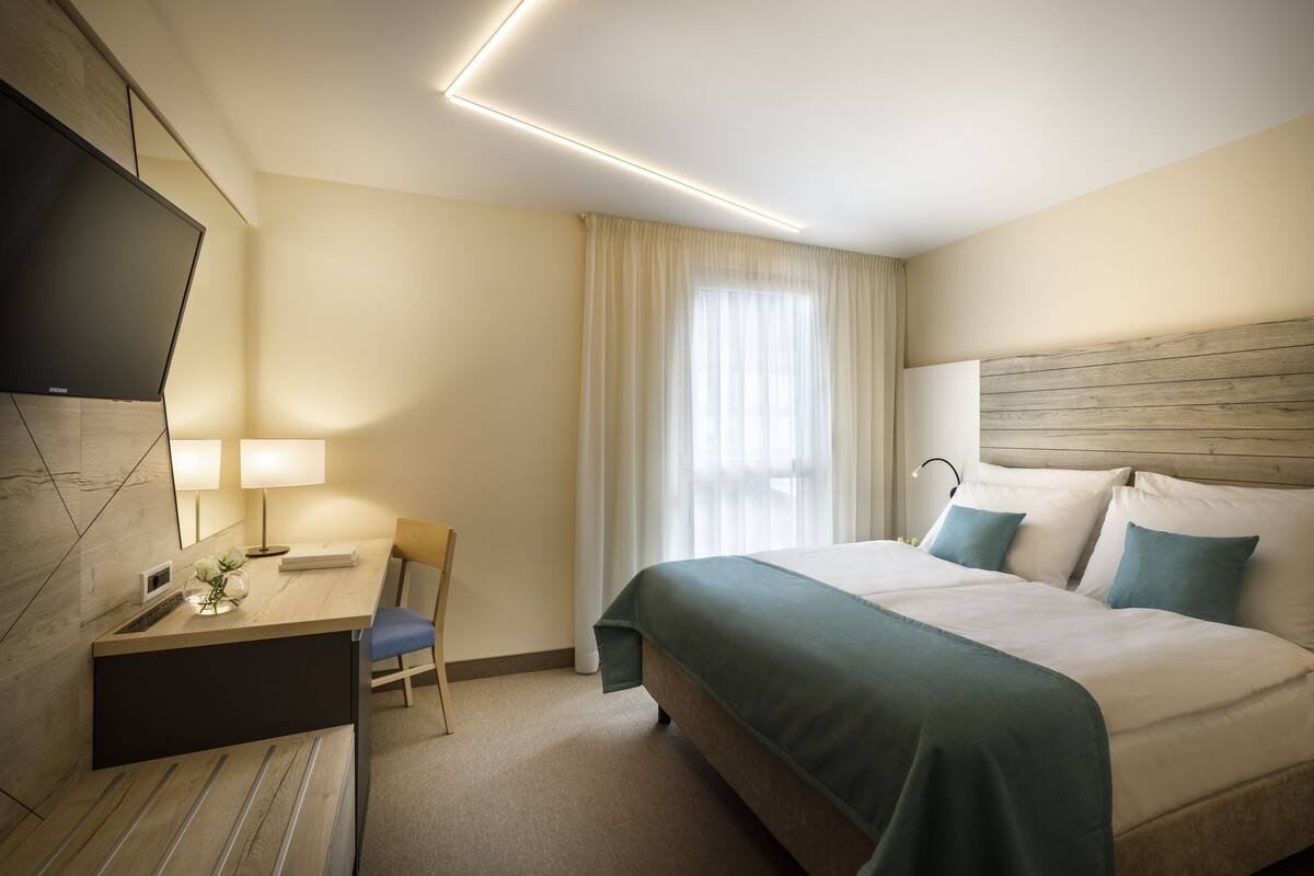 A hotel room with a bed, a desk with a chair, a television, and a window with curtains.