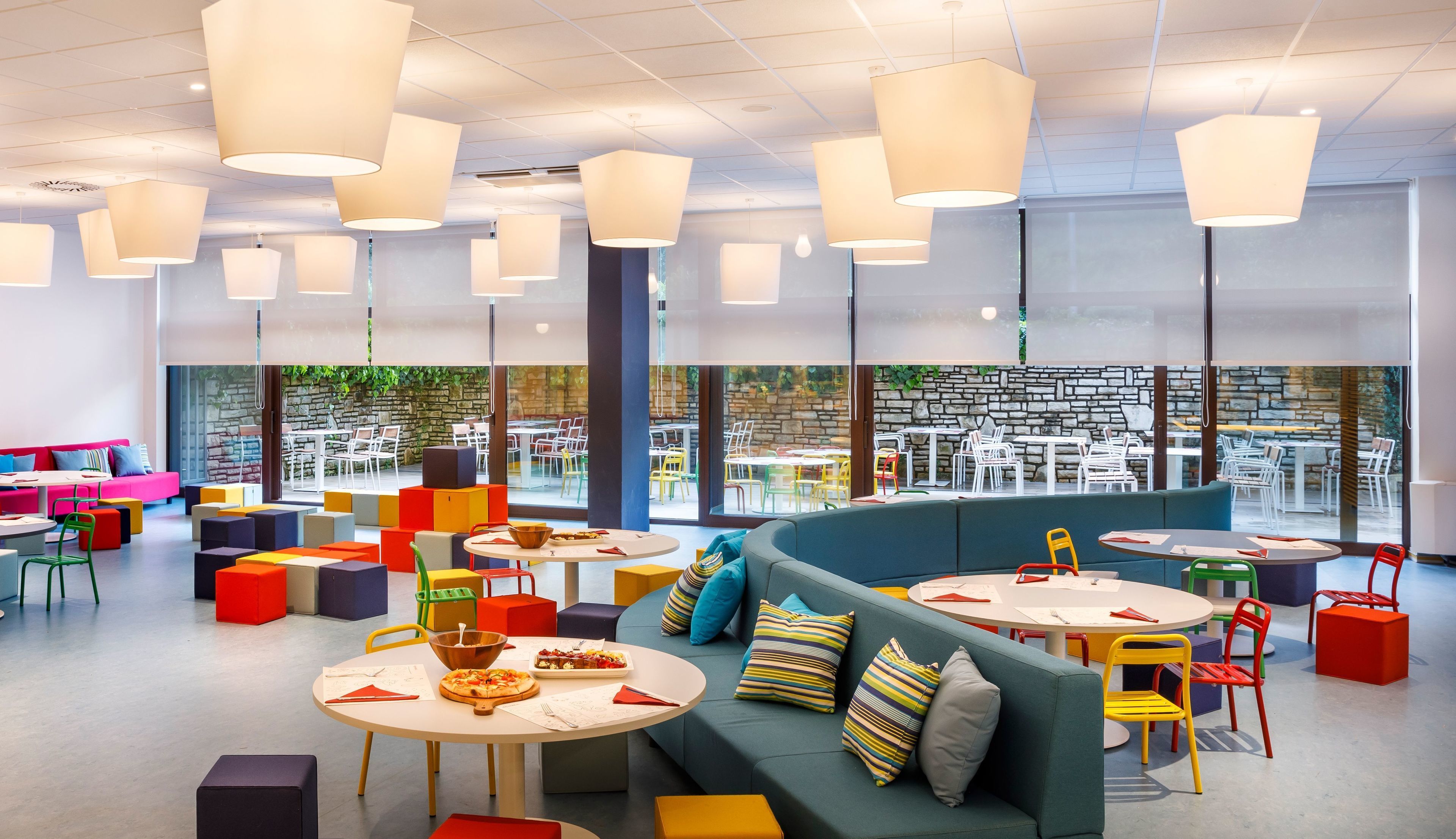 Un espace de restauration intérieur moderne, doté de sièges et de tables aux couleurs vives, de grandes fenêtres et d’un mur en pierre en arrière-plan.