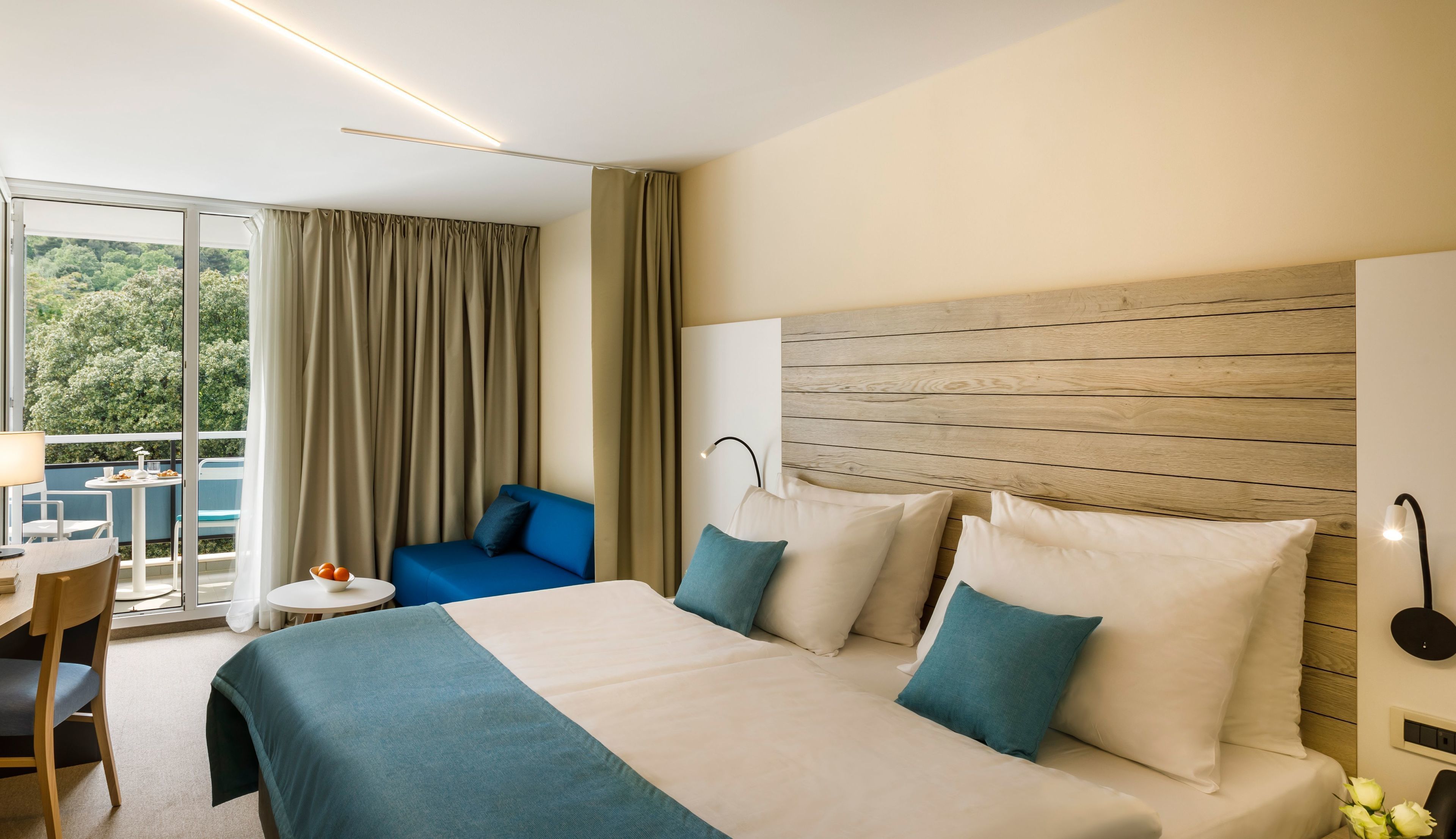 A hotel room with a large bed, a blue sofa, a small table with a lamp, and a view of greenery through the window.