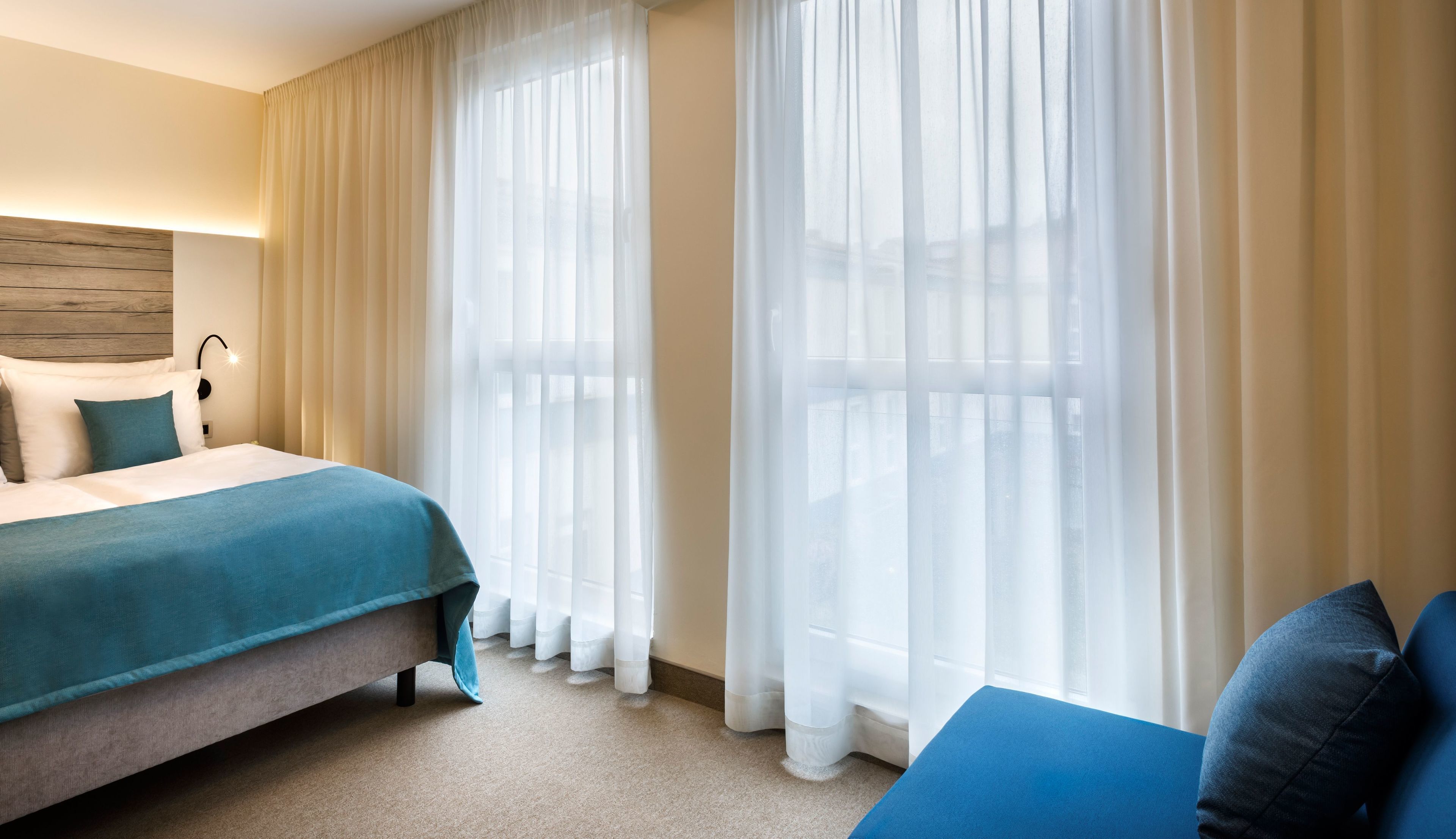Una habitación de hotel con cama, sofá y ventanas amplias con cortinas translúcidas.