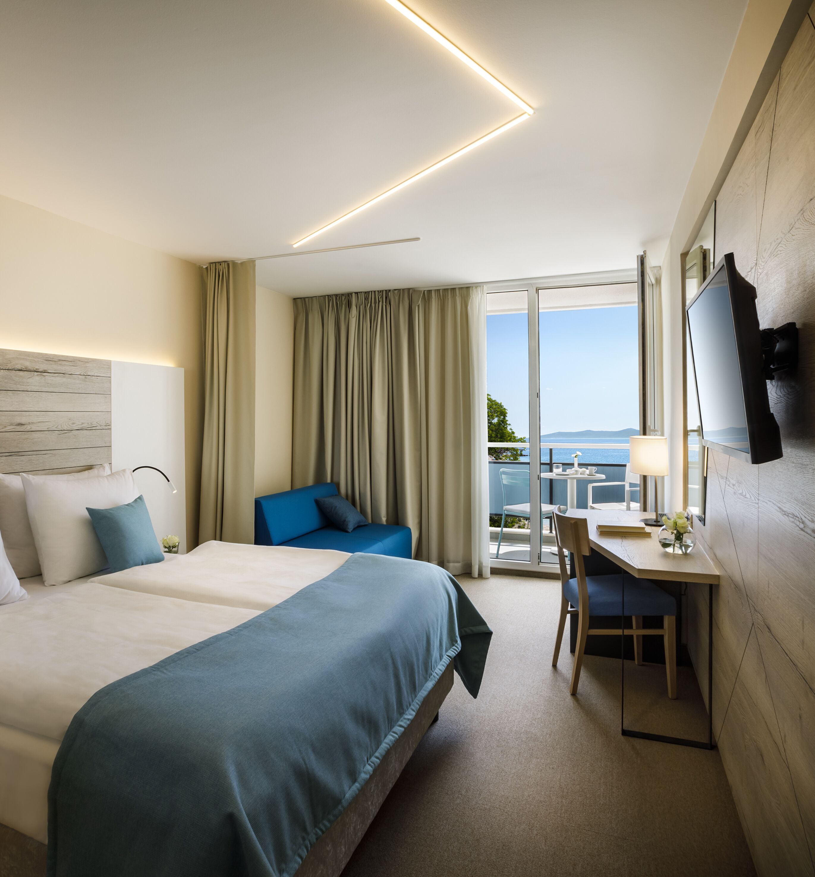 Habitación de hotel con una cama grande, un escritorio con silla, un televisor montado en la pared y un balcón con vista al mar.