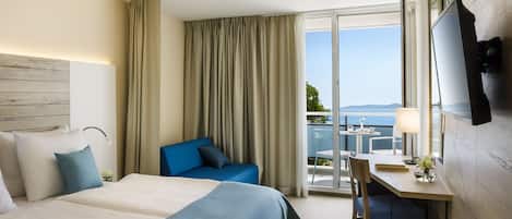 A hotel room with a large bed, a desk with a chair, a TV mounted on the wall, and a balcony with a view of the sea.