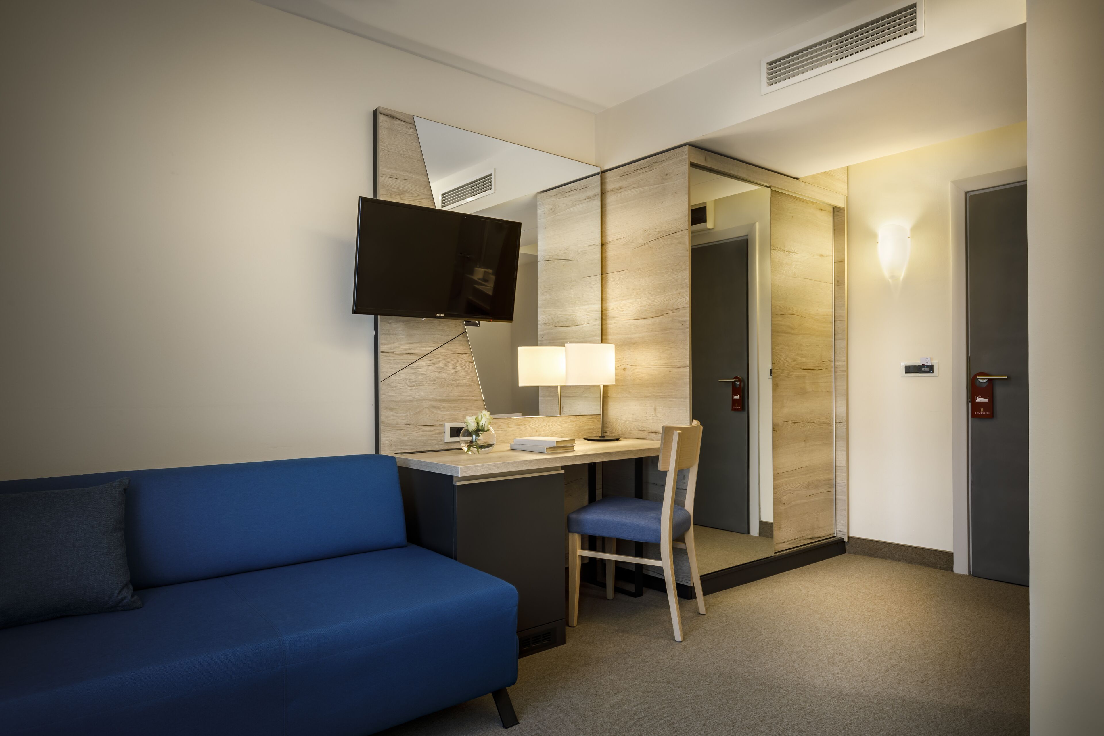 A hotel room with a blue sofa, a desk with a chair, a flat-screen TV, a lamp, and a mirror.