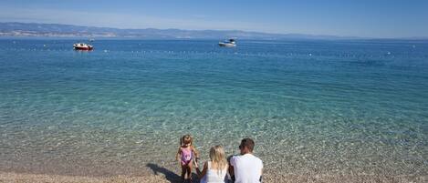 A pebbled beach with clear water and boats in the distance.