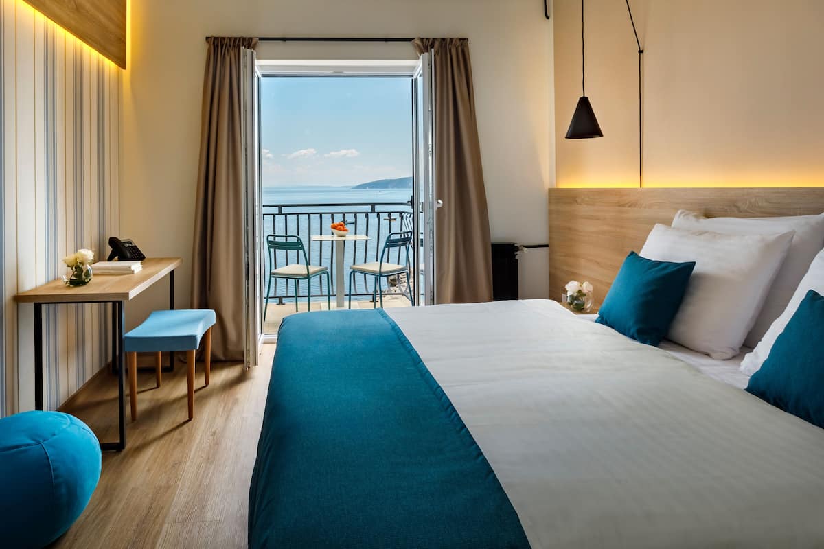 Een hotelkamer met een bed, een bureau, een stoel, een klein balkon met een tafel en stoelen, en uitzicht op de zee.