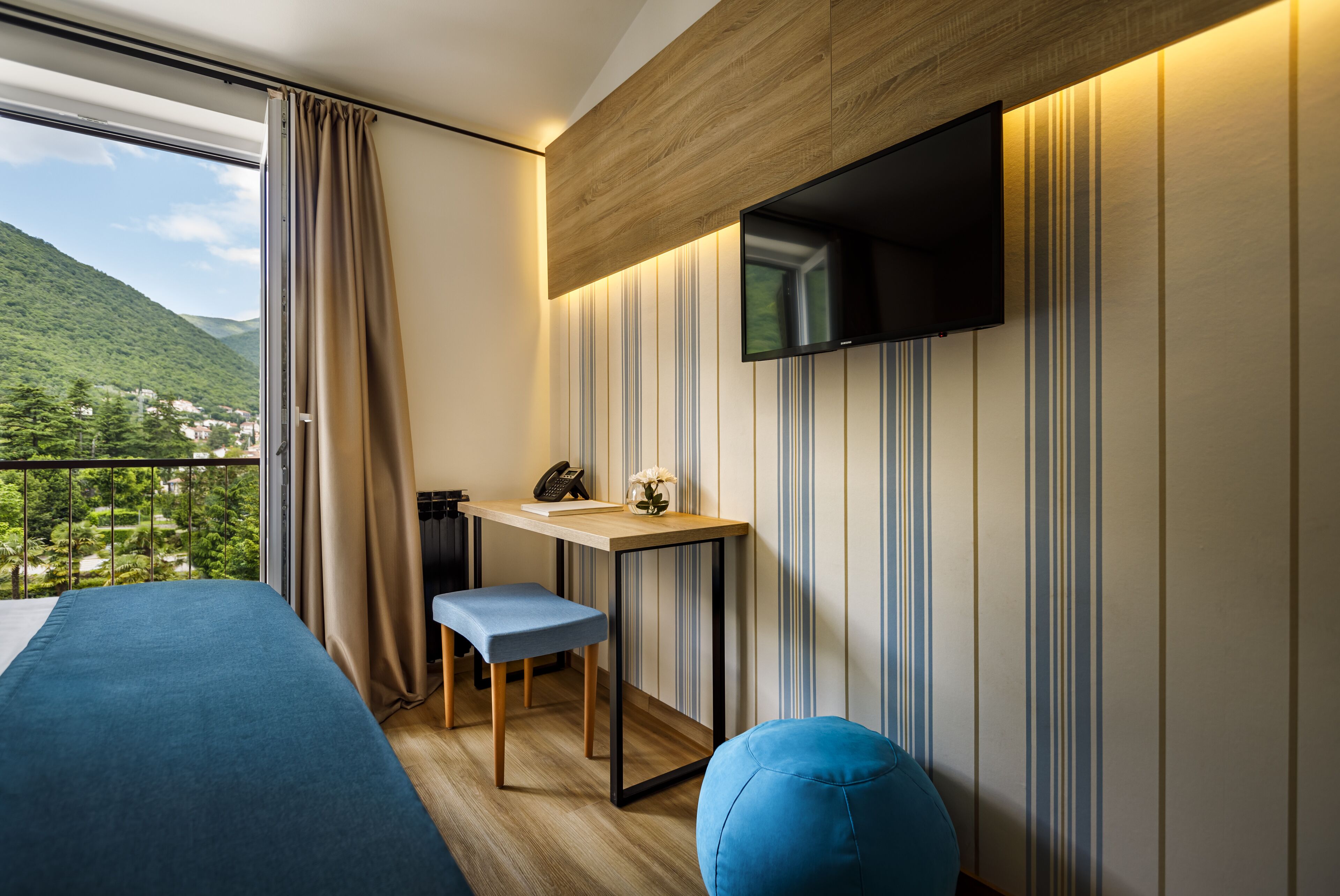 A modern hotel room with a large window, a bed, a desk with a chair, a television, and a view of a mountainous landscape.