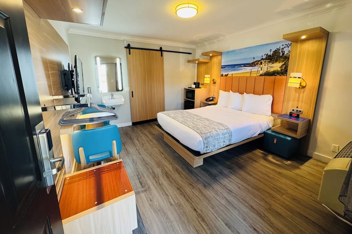 A hotel room with a bed, a desk with a chair, a sink, and a painting of a beach scene.