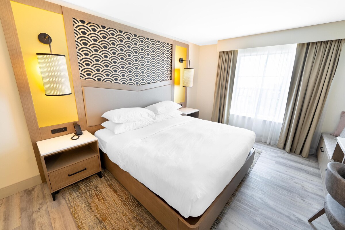 A hotel room with a large bed, a nightstand, a lamp, and a view of the outdoors through a window with curtains.
