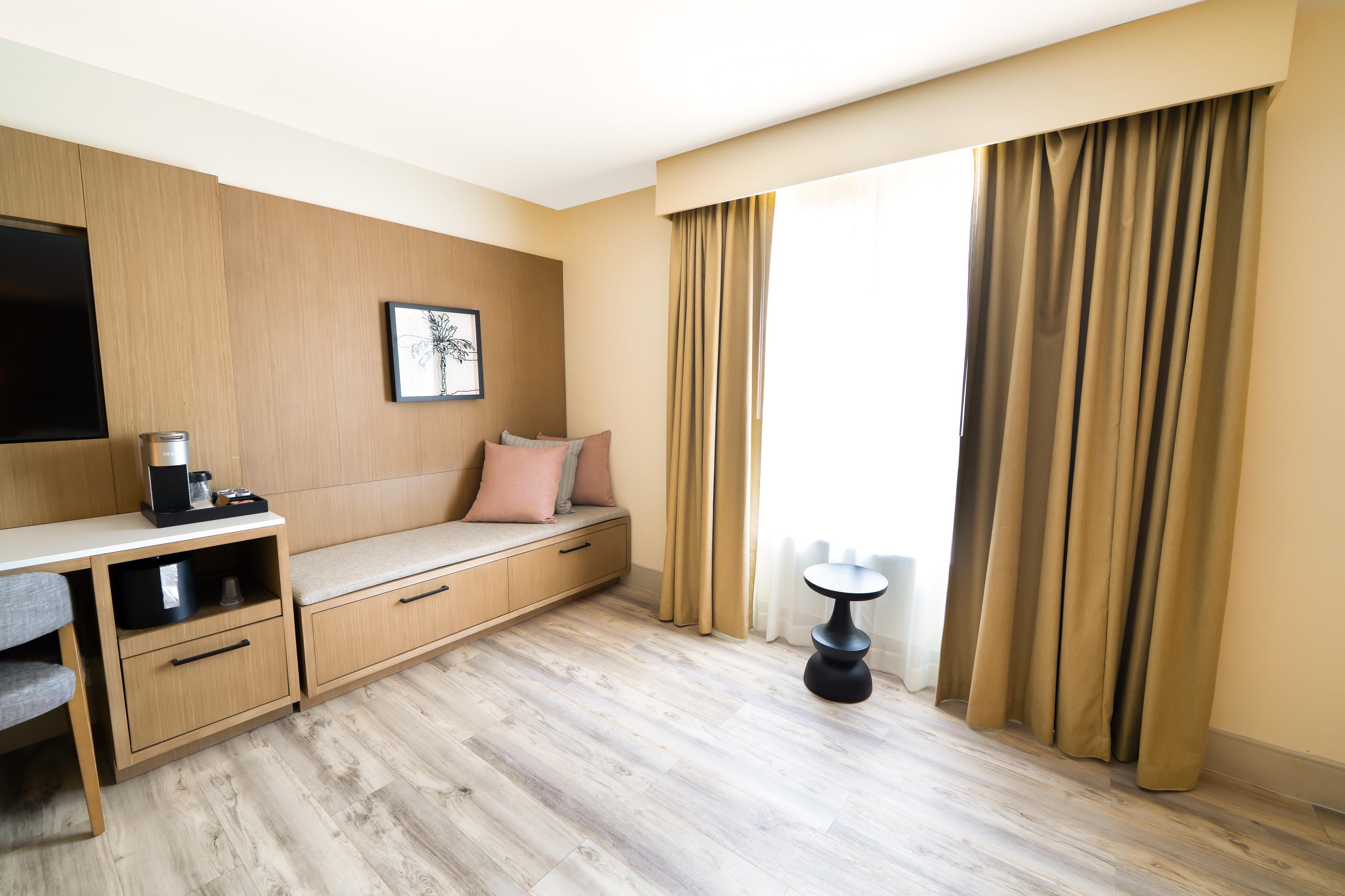 A hotel room with a wooden desk, a bench with cushions, a coffee machine, a framed picture on the wall, and a window with curtains.