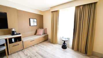 A hotel room with a wooden desk, a bench with cushions, a coffee machine, a framed picture on the wall, and a window with curtains.