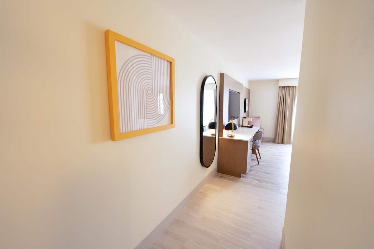 A hotel room with a wooden floor, a desk with a chair, a large mirror, and a framed artwork on the wall.