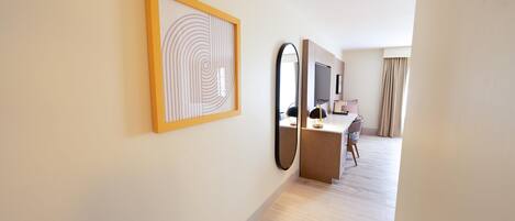 A hotel room with a wooden floor, a desk with a chair, a large mirror, and a framed artwork on the wall.