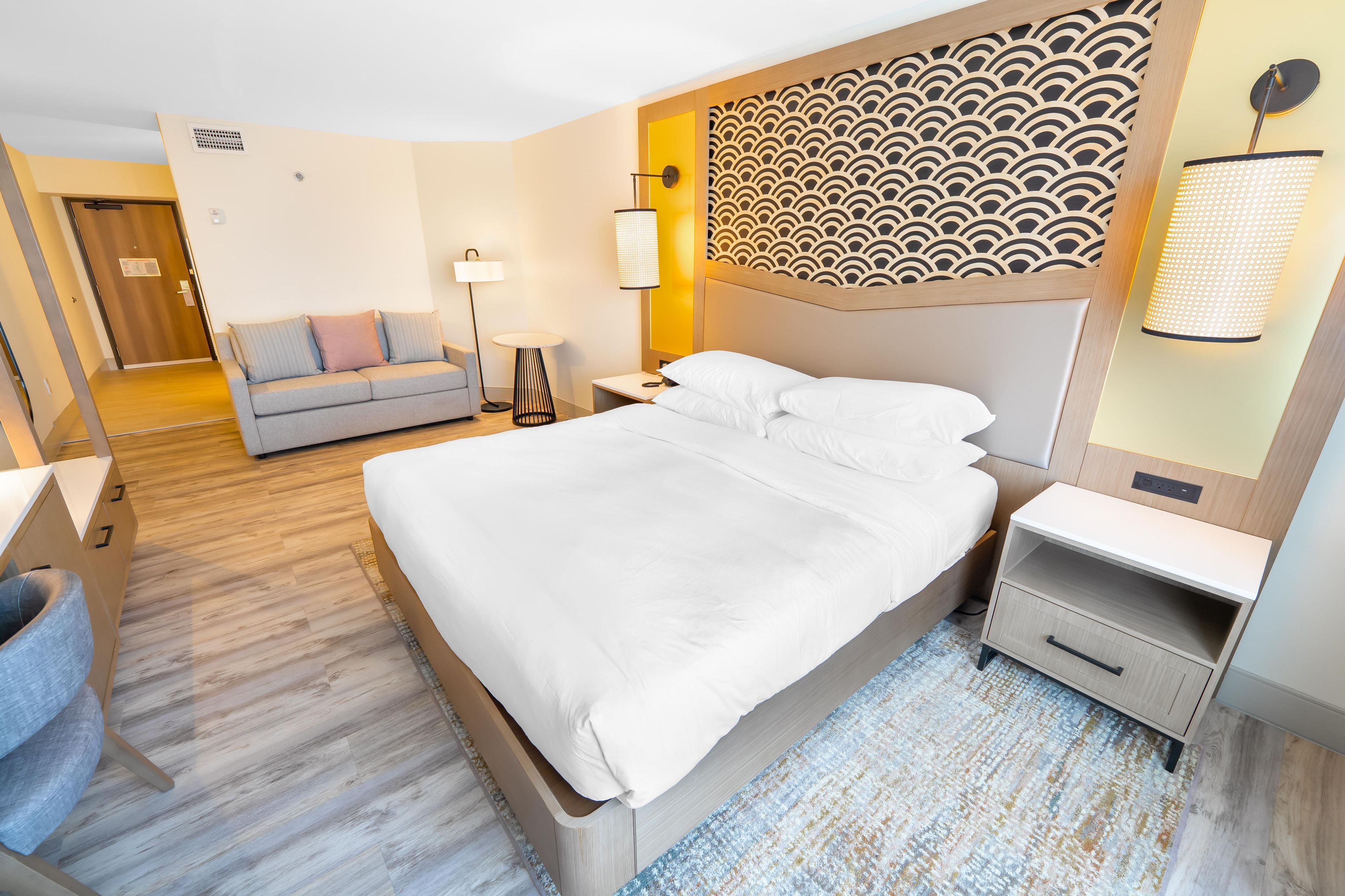Una habitación de hotel con una cama grande, una mesita de noche, una lámpara, un sofá y un papel tapiz estampado.