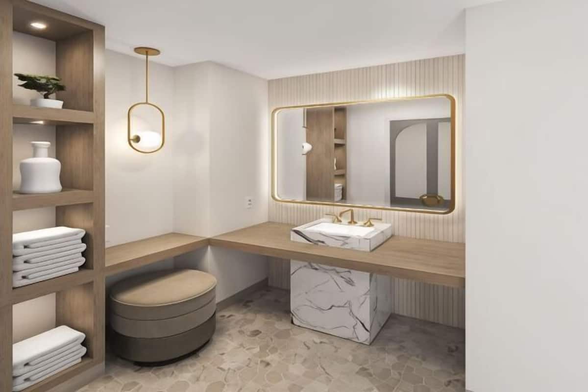 A modern bathroom with a wooden vanity, a round stool, a marble base, and a large mirror with a gold frame.
