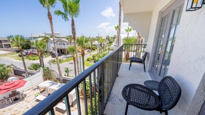 A balcony with two black wicker chairs, a red umbrella, and a view of a resort area with palm trees and buildings. - Castillo Real St. Augustine Beach,Tapestry Collection Hilton (St. Augustine)