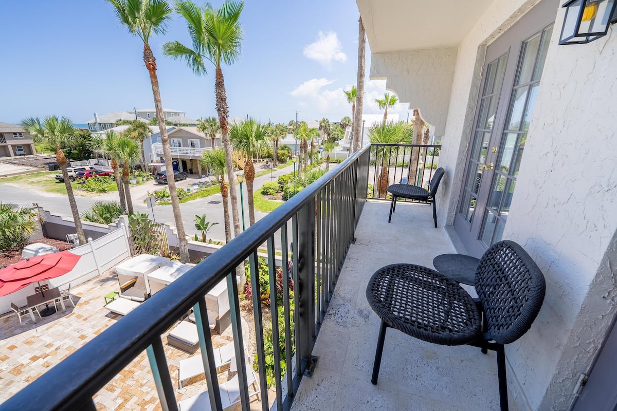A balcony with two black wicker chairs, a red umbrella, and a view of a resort area with palm trees and buildings.