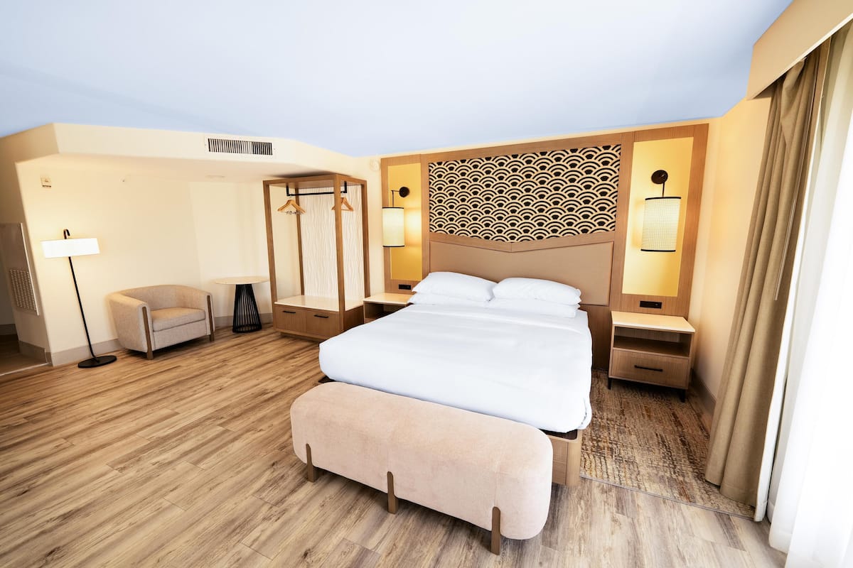 A hotel room with a large bed, a bedside table, a lamp, a chair, a wardrobe, and a patterned headboard.