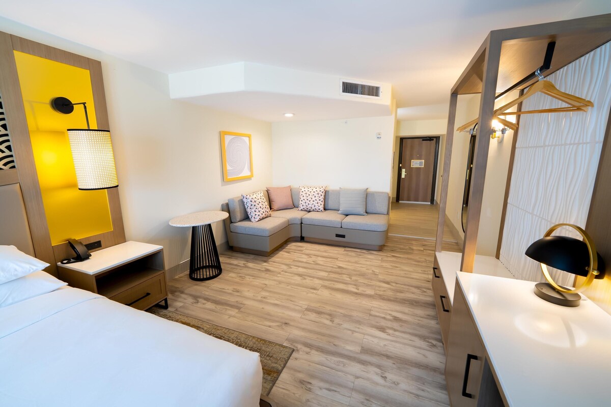A modern hotel room with a bed, a sofa, a desk, and a mirror.