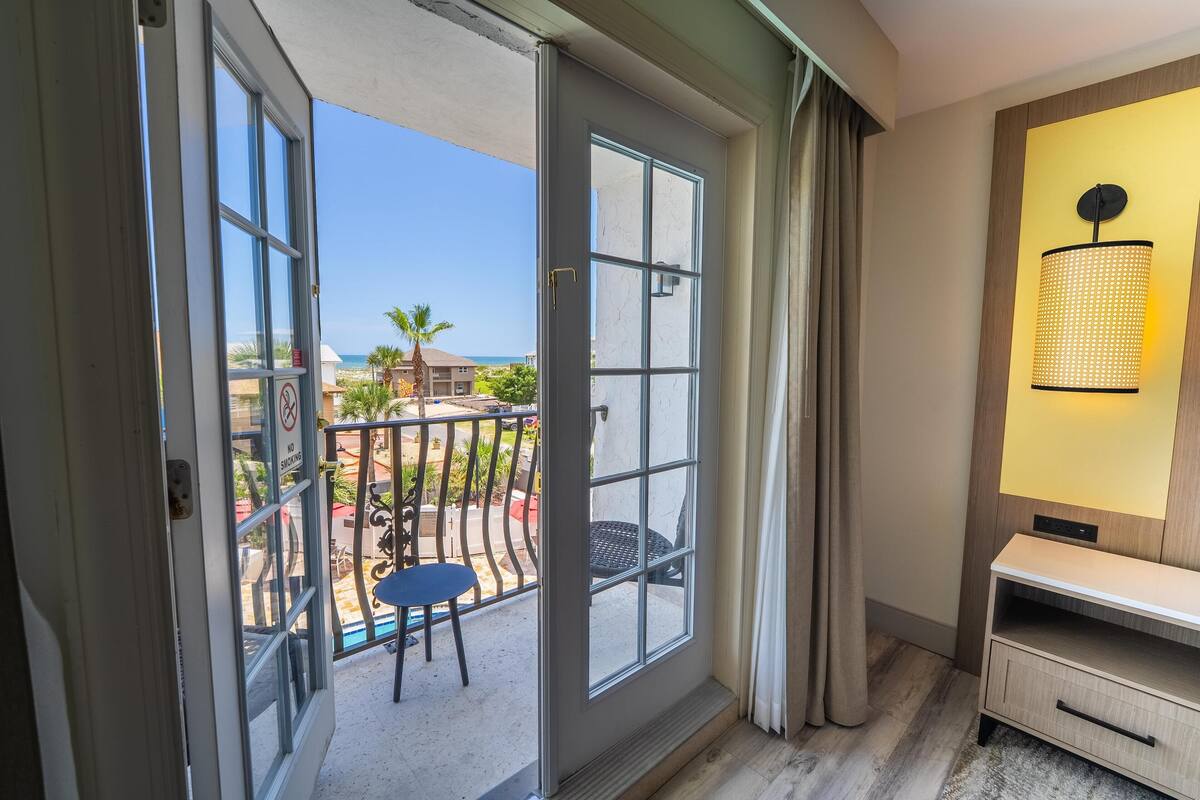 A room with a sliding glass door leading to a balcony with outdoor furniture and a view of the ocean.