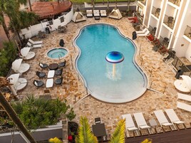 Una piscina con lettini e vasca idromassaggio, circondata da un solarium piastrellato e palme.
