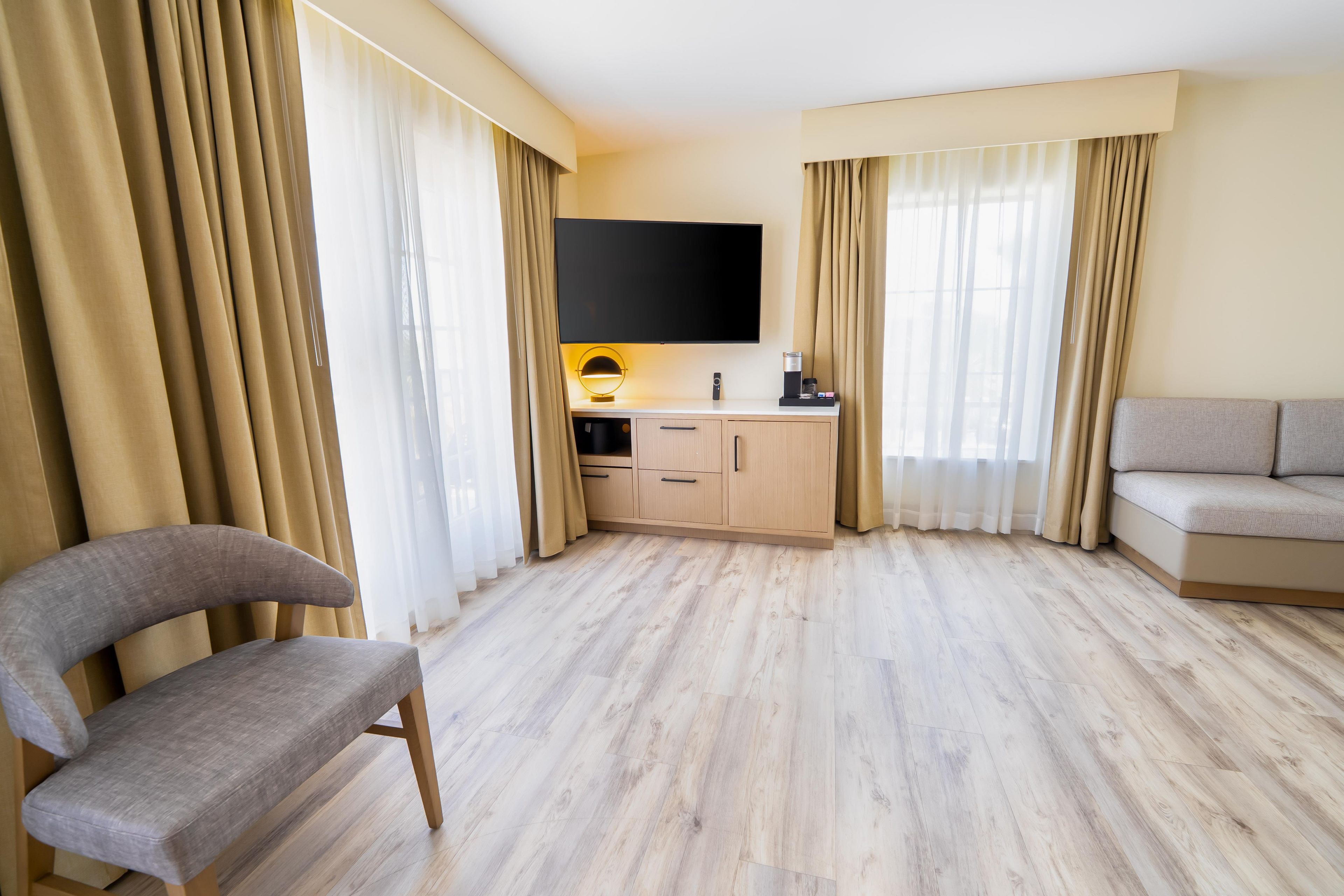 Habitación de hotel con televisor de pantalla plana sobre un escritorio de madera, un sofá gris y un sillón gris.