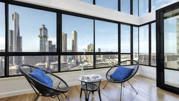 A high-rise room with large windows, two black wireframe chairs with blue cushions, a small table with a white vase and magazine, and a wooden floor.