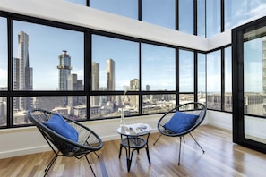 Habitación en un rascacielos con ventanas amplias, dos sillas de alambre negras con cojines azules, una mesita con un jarrón blanco y una revista, y suelo de madera.