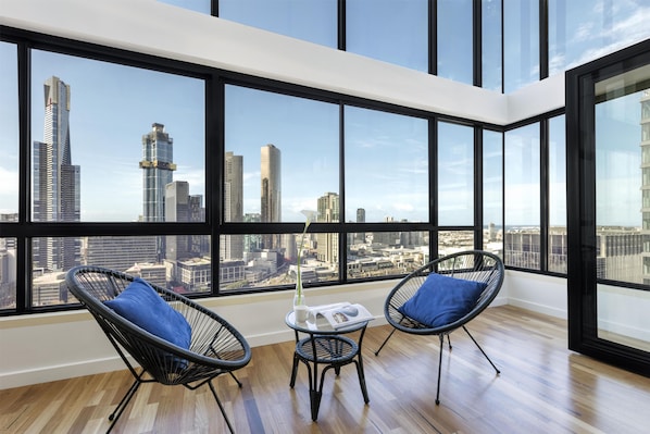 A high-rise room with large windows, two black wireframe chairs with blue cushions, a small table with a white vase and magazine, and a wooden floor.