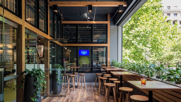 An interior with wooden flooring, multiple tables and stools, and large windows with a view of greenery outside.